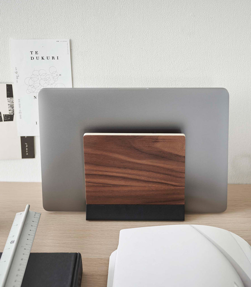 View 11 - A small dark wood laptop stand with a black metal base stores a closed laptop on a wooden desk.