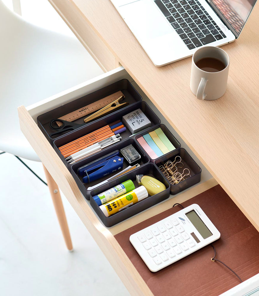 View 63 - Three different sizes of stackable clear organizers in black —small, medium, and large—are neatly placed in a desk drawer. Made from transparent material, they accommodate stationery items like pens, paper clips, and sticky notes, showcasing their clarity and stackability.