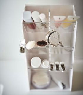 An upper view of a white rectangular resin cosmetics organizer on a white bathroom counter. It has three deep transparent trays that sit diagonally with adjustable transparent dividers placed in the middle of each tray. The middle tray is in focus and holds a set of cosmetic brushes and an eyelash curler. view 7