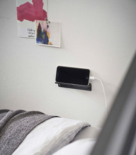An iPhone with a charger cord plugged in is mounted on a wall above a bed setup. The screen is facing outward and the phone is held up on its side. view 15