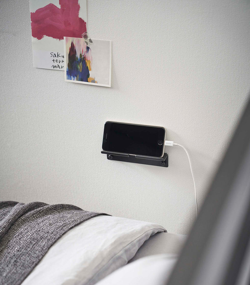 An iPhone with a charger cord plugged in is mounted on a wall above a bed setup. The screen is facing outward and the phone is held up on its side.