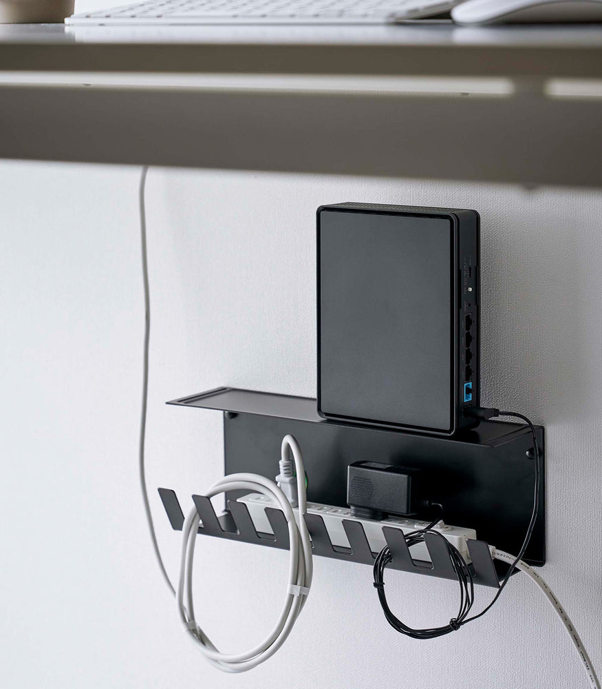 View 12 - Under-Desk Cable Organizer in black by Yamazaki Home mounted on a wall holding a power strip and a router.