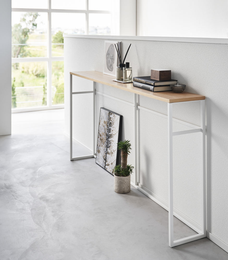 View 3 - Diagonal view of the Long Console Table by Yamazaki Home in white placed by a wall with decorative items.