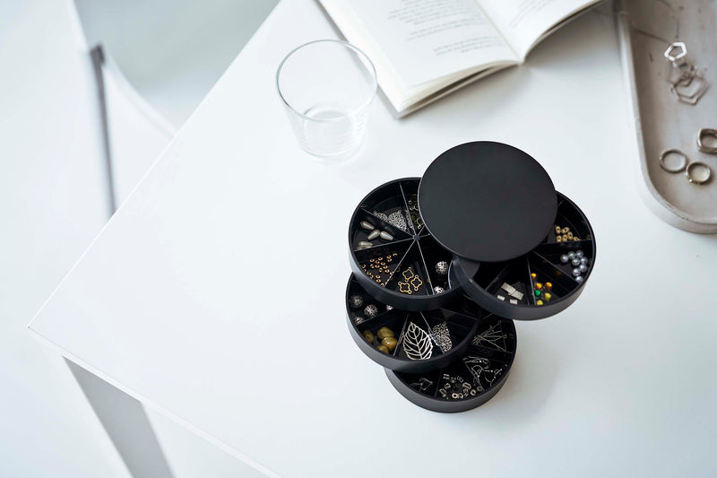 A bird’s-eye view of the corner of a white table. On the table are a cylinder-shaped black four-tier swivel accessory holder, a clear drinking glass, and the corner of an opened book. The accessory holder’s tiers and lid are swiveled opened so the inside contents can be seen. Each tier of the accessory holder is divided into seven separate compartments in a pie-chart fashion. Each compartment holds various beads and other jewelry-making parts.