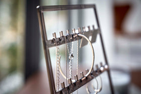 A detailed view of an acrylic translucent mauve earring holder. The holder has upward pointed hooks and slots placed in an interchangeable pattern. Hanging from the hooks are chained necklaces, and in the slots are various earrings. view 12