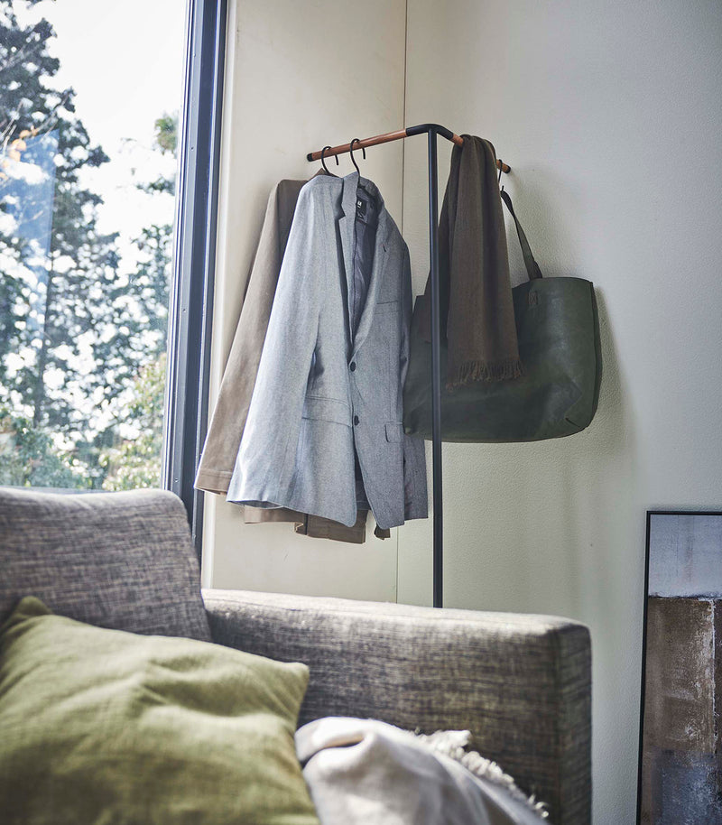 Side view of Yamazaki black Corner Leaning Coat Hanger in a the corner of a room with clothing hung on it