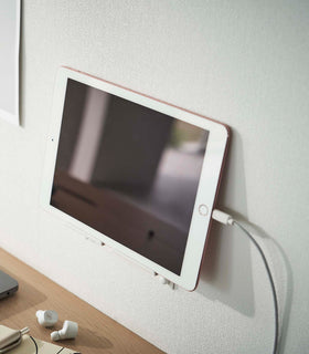 A tablet with a charger cord plugged in is mounted on a wall above a desk setup. The screen is facing outward and the tablet is held up on its side. view 3