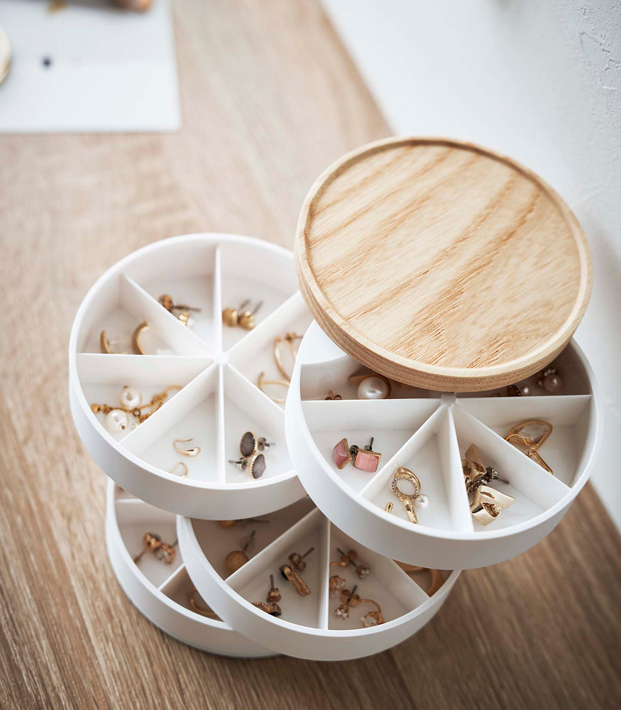 View 10 - A four-tier swivel accessory organizer with a light-colored wooden lid is displayed in an all-white room. The accessory holder’s tiers and lid are swiveled opened so the inside contents can be seen, each tier has seven individual compartments formed in a pie shape. Each individual compartment of the tiers holds a pair of earrings.