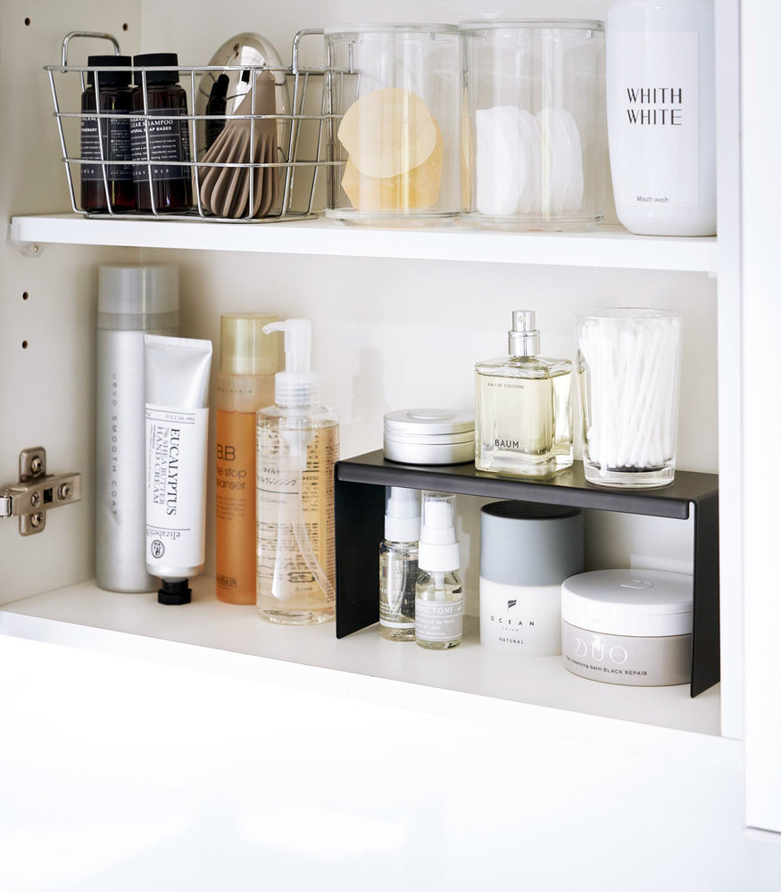 View 9 - Open medicine cabinet showing black countertop riser on bottom shelf. Upper level of the riser holds perfume and hair products, while skincare and small sprays are stored on the bottom.