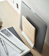 A side-view profile of a narrow wooden stand with a white metal base positioned by books on top of a desk.