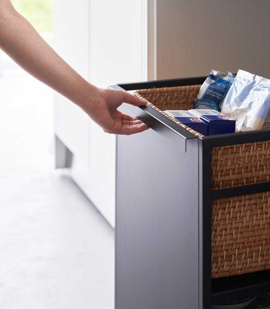 View 15 - Two-level black steel storage cart with casters is shown in an opening of a recessed cabinet. A model demonstrates its mobility by pulling on the handle.