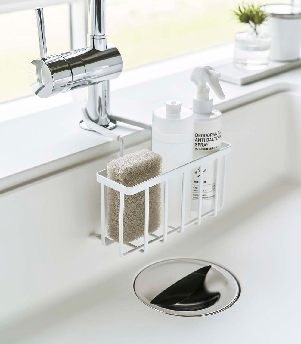 White Faucet-Hanging Sponge Caddy attached to the kitchen sink faucet and holding sponges and cleaning supplies by Yamazaki Home.