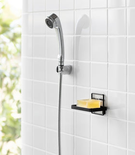Yamazaki Home's black Traceless Adhesive Soap Tray in a shower, holding a bar of soap next to a silver shower hose on tiled wall. view 10