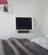 A tablet with a charger cord plugged in is mounted on a wall above a bed setup. The screen is facing outward and the tablet is held up on its side.