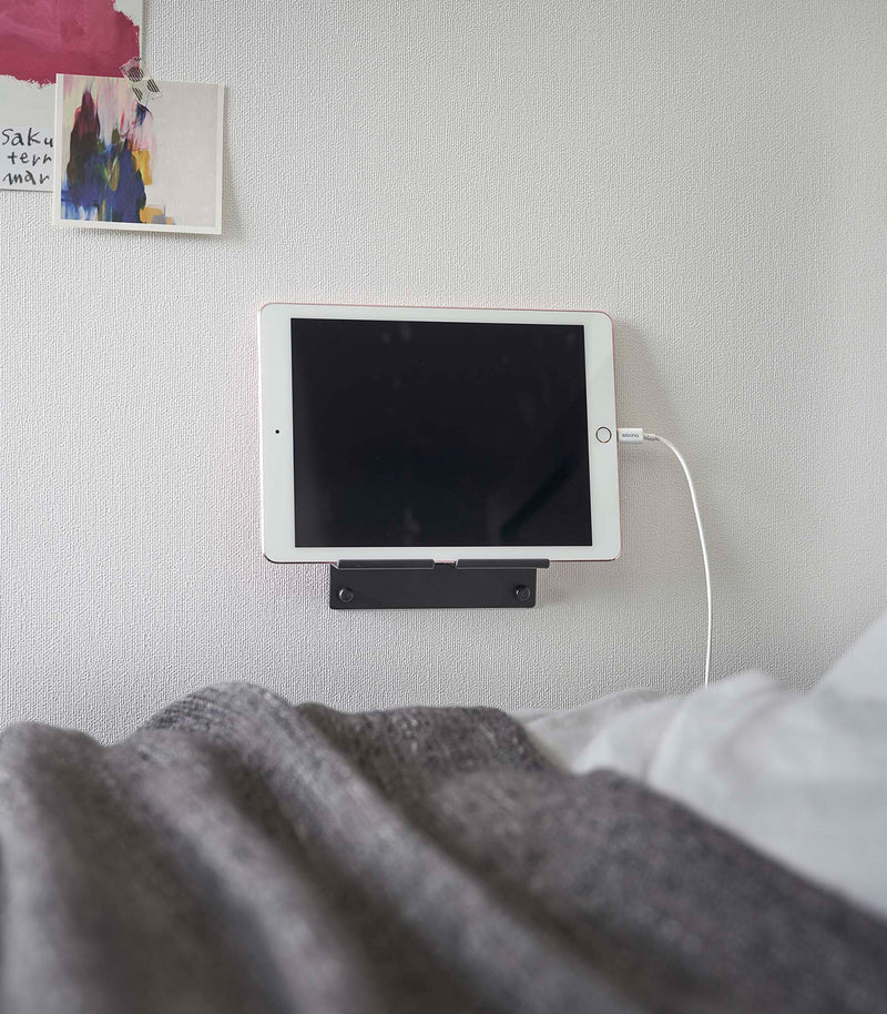 A tablet with a charger cord plugged in is mounted on a wall above a bed setup. The screen is facing outward and the tablet is held up on its side.