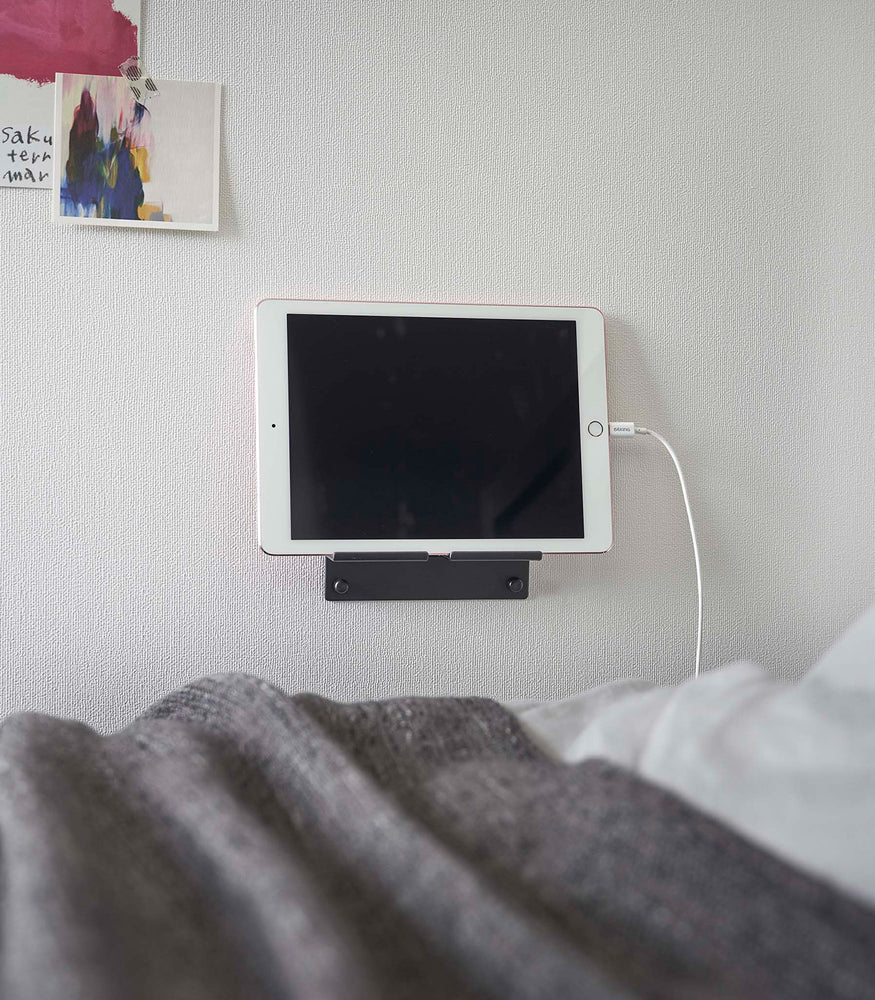 View 16 - A tablet with a charger cord plugged in is mounted on a wall above a bed setup. The screen is facing outward and the tablet is held up on its side.