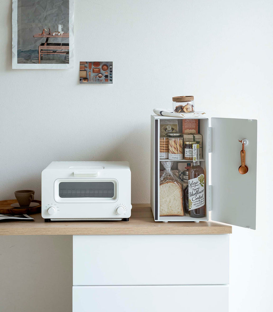 View 10 - A vertical white metal breadbox is seen on a wooden kitchen counter next to a white microwave oven. The breadbox’s door is swung open to the right and bread, other grains, and a bottle of wine are seen inside the box. A magnetic stop is seen opposite the open door. On the open door is a white hook holding brown plastic measurement spoons. On top of the box is a folded towel and plastic container of cookies.