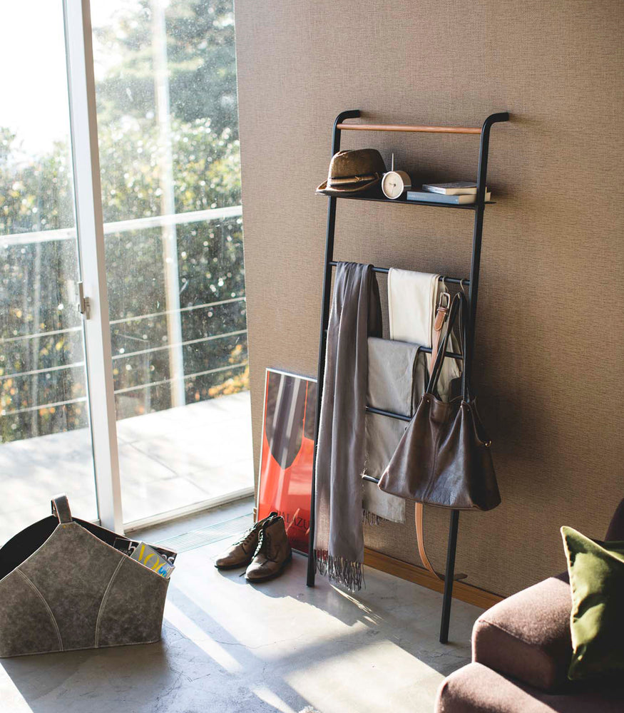 View 20 - Black Leaning Ladder Rack with Shelf holding clothing items, books, and a clock in bedroom by Yamazaki Home.