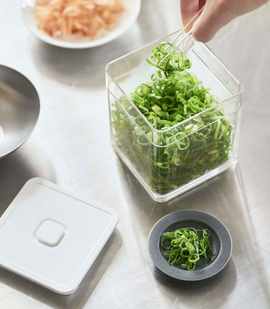 View 4 - Aerial view of person using tongs to grab scallions out of white Vacuum-Sealing Food Container by Yamazaki Home.