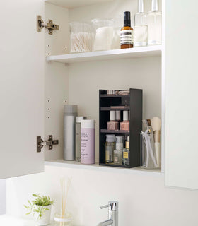 A white medicine cabinet is open to display the inside contents. Sunlight is focused on the right upper corner. Below is a bathroom sink. On the sinks ledge is a plant and oil diffuser. A white shelf divides the cabinet. The bottom section features a cosmetic organizer. view 30