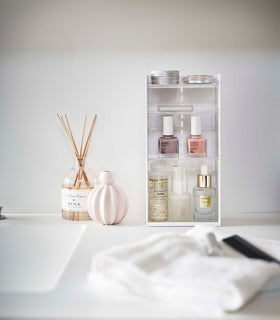 A white rectangular cosmetics organizer sits on a white bathroom counter. The organizer has three transparent shelves with upward facing lips to prevent products from falling-out. view 3