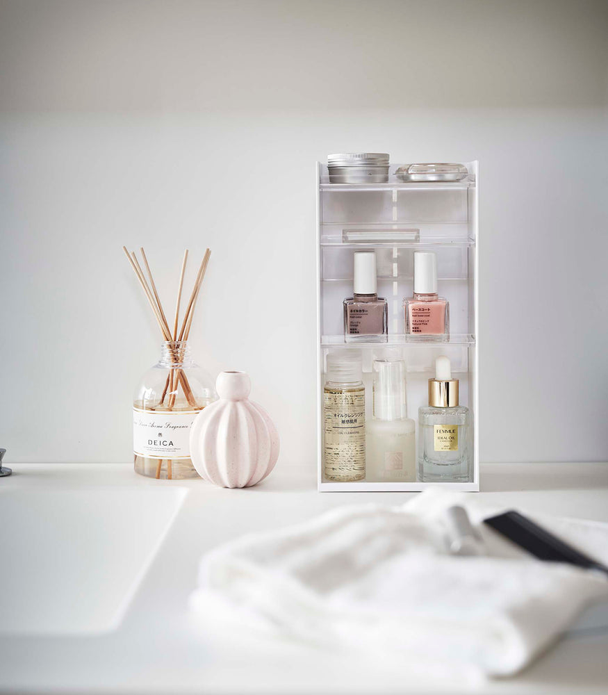 View 3 - A white rectangular cosmetics organizer sits on a white bathroom counter. The organizer has three transparent shelves with upward facing lips to prevent products from falling-out.