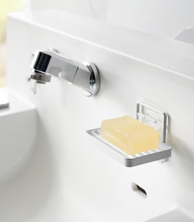 Yamazaki Home's white Traceless Adhesive Soap Tray mounted on a bathroom wall, holding a bar of yellow soap next to a chrome faucet. view 2