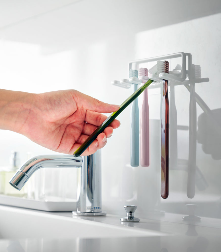 View 5 - Yamazaki Home's Traceless Adhesive Toothbrush Holder, white, mounted on a bathroom wall with four colorful toothbrushes and a chrome faucet.