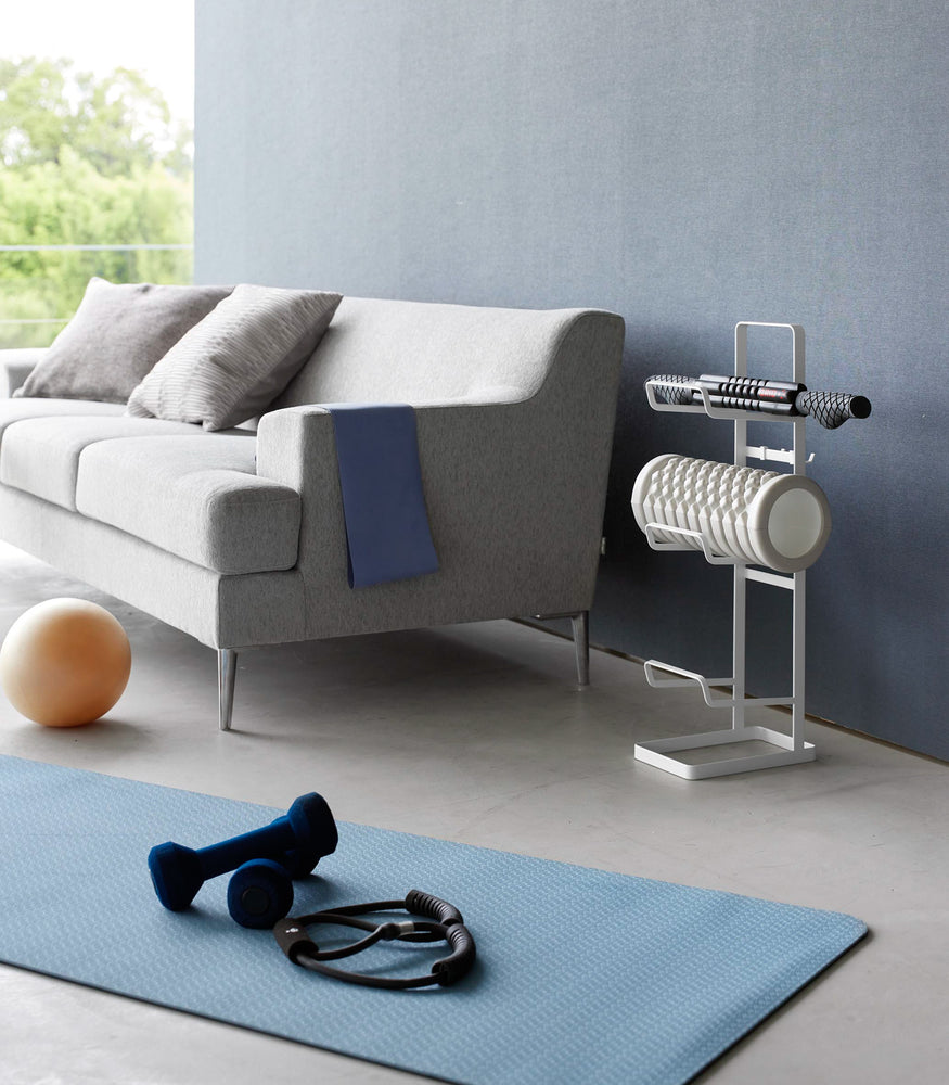 View 3 - Yamazaki Home White Fitness Equipment Storage Rack holding foam rollers next to sofa and sports equipment.