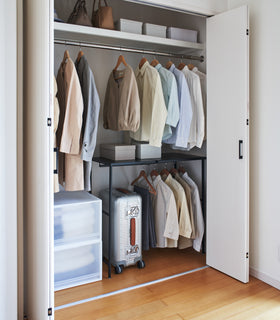 Black Yamazaki Home Expandable Suitcase Rack in a closet with clothes hung and a suitcase underneath view 15