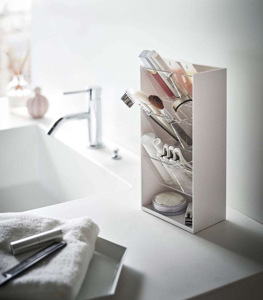 View 2 - An angled view of a white rectangular resin cosmetics organizer on a white bathroom counter. It has an open face and top and three deep transparent trays that sit diagonally with adjustable transparent dividers placed in the middle of each tray for easy visibility.