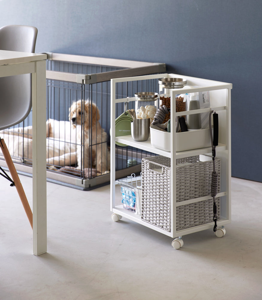 View 4 - Two-level white steel storage cart with casters is shown next to a dog crate. Cart houses various dog supplies and treat storage on the upper shelf, bottom shelf holds two storage bins. Hooks are installed on both sides, one holds a collapsible water bowl, the other a leash. Solid back wall of the cart demonstrates that the items can be hidden when flipped.