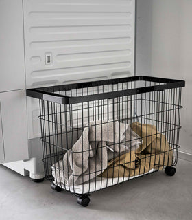 Rolling Wire Basket by Yamazaki Home in black stowed next to a laundry machine with several towels inside. view 14