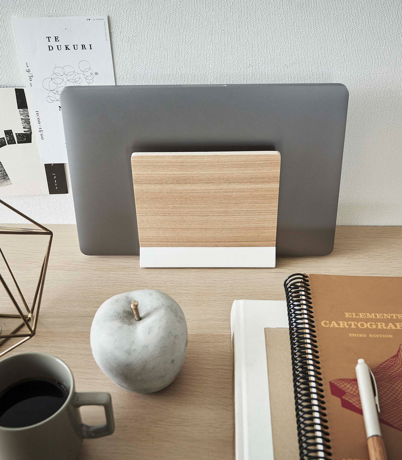 A small light-colored wood laptop stand with a white metal base stores a closed laptop on a wooden desk.