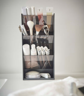 A front view of a black rectangular resin cosmetics organizer on a white bathroom counter. It has three deep adjustable black transparent trays that sit diagonally with matching dividers placed in the middle of each tray. view 8
