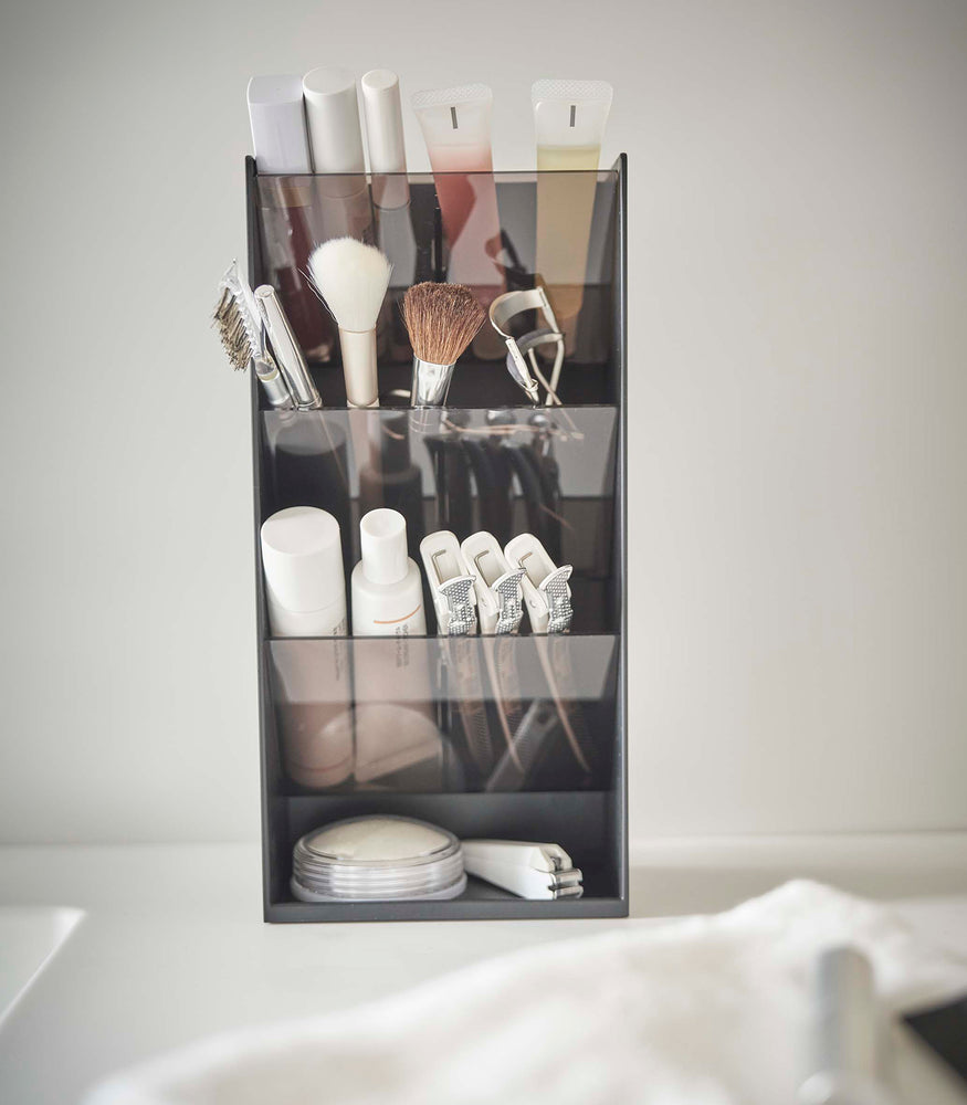 View 8 - A front view of a black rectangular resin cosmetics organizer on a white bathroom counter. It has three deep adjustable black transparent trays that sit diagonally with matching dividers placed in the middle of each tray.