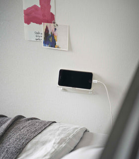 An iPhone with a charger cord plugged in is mounted on a wall above a bed setup. The screen is facing outward and the phone is held up on its side. view 8