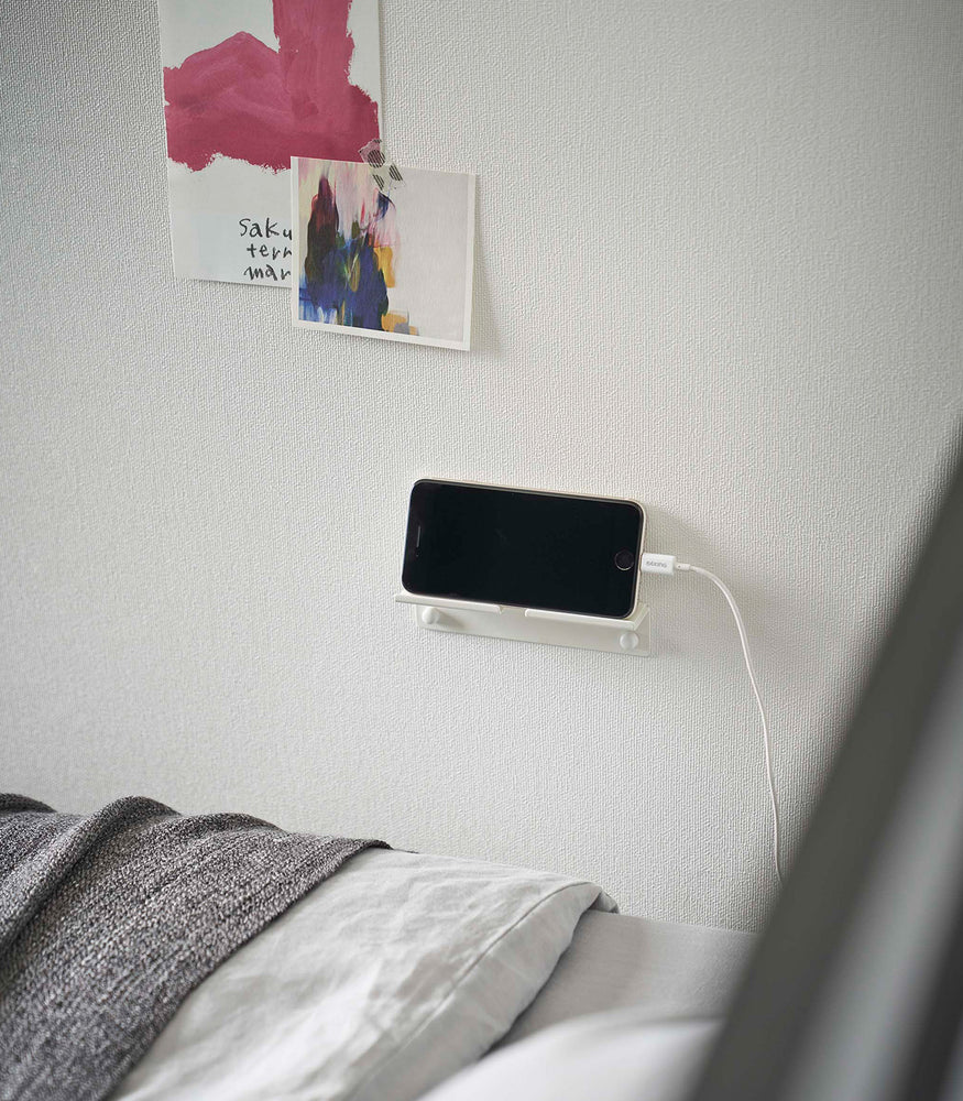 View 8 - An iPhone with a charger cord plugged in is mounted on a wall above a bed setup. The screen is facing outward and the phone is held up on its side.