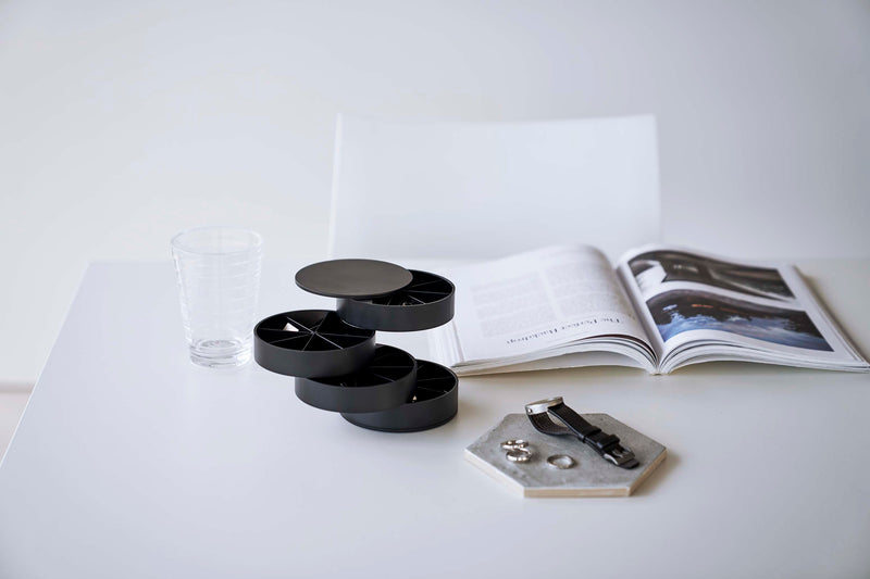 A front facing view of a white table with a white chair pushed in. On the surface of the table are a cylinder-shaped black four-tier swivel accessory holder, a clear drinking glass, and an opened book facing the direction of the chair. Each tier of the accessory holder is divided into seven separate compartments in a pie-chart fashion.