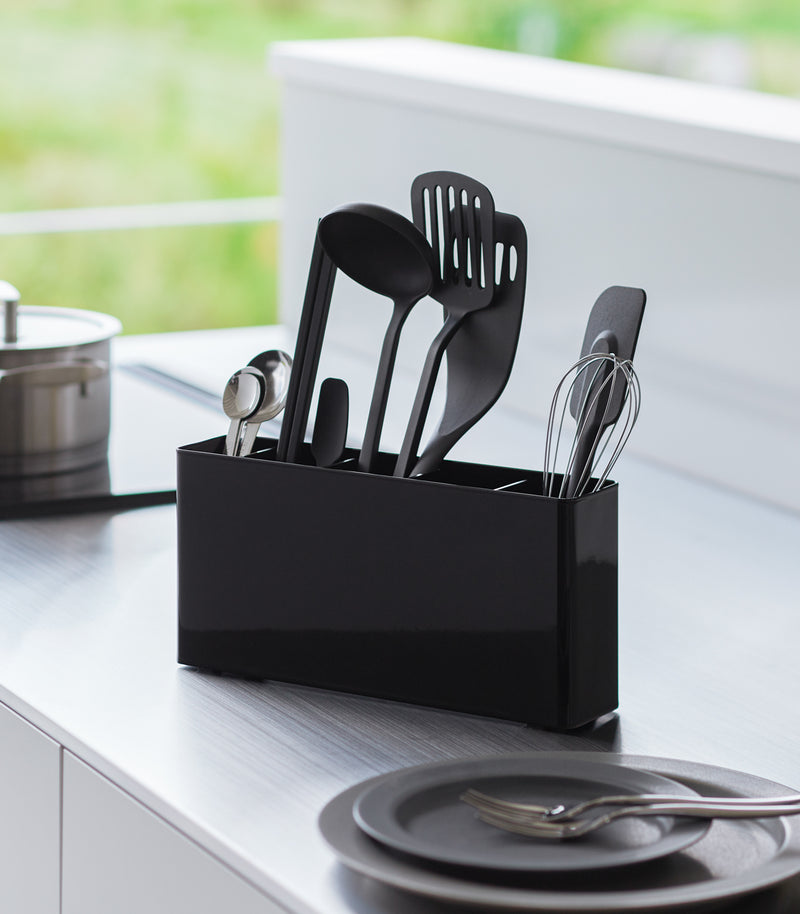 Black Utensil & Thin Cutting Board Holder by Yamazaki Home on a kitchen counter, holding various utensils and a cutting board.