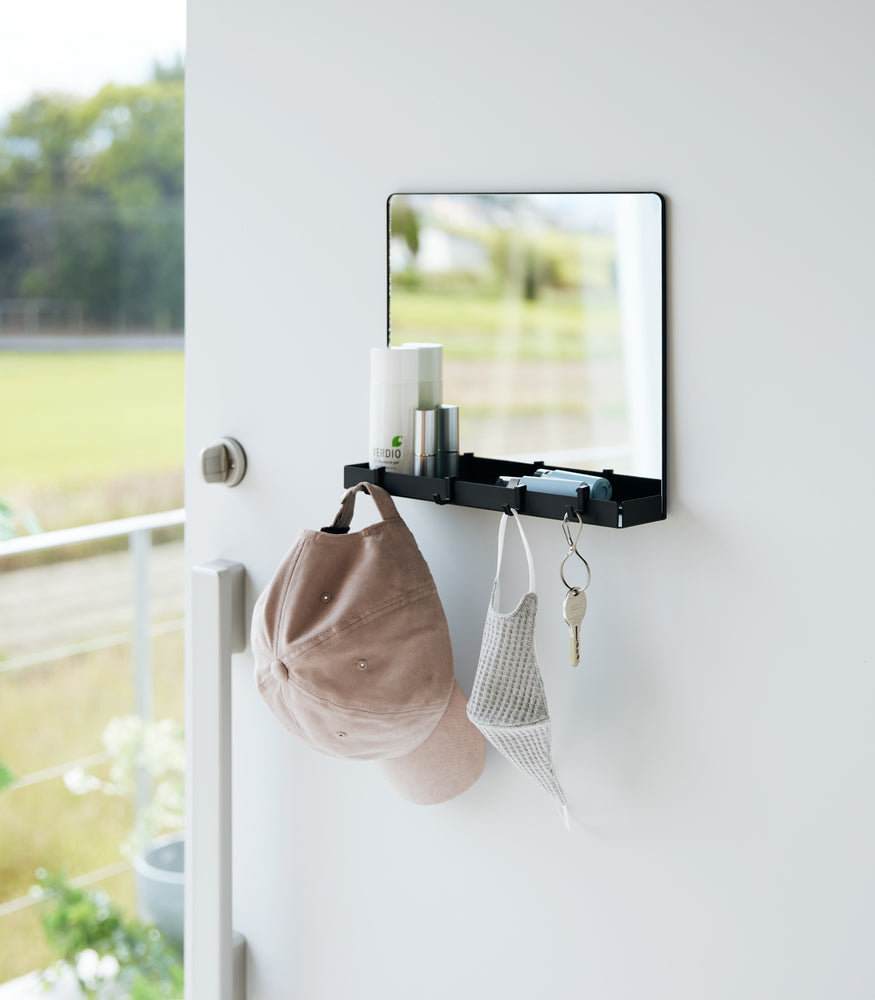 View 14 - A black Yamazaki Magnetic Mirror With Storage Rack attached to a door with a hanging hat, mask, and key with a view outside.