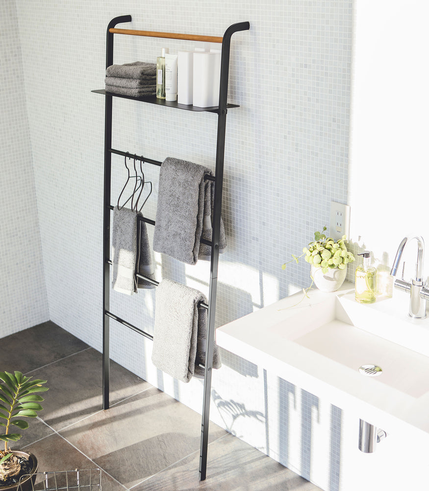View 21 - Black Leaning Ladder Rack with Shelf holding towels, hangers, and skin care products in bathroom by Yamazaki Home.