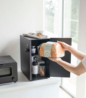 An angled view of a person’s arms putting bread into a black metal vertical breadbox placed on top of a white kitchen counter. The door is swung open from the right and a magnetic strip is seen opposite of its position. Coffee Syrup, cinnamon sticks in a clear container, and other pantry items are seen in the open breadbox. A magnetic hook is attached to its side. On top is a folded tea towel and clear package of treats. Slightly off frame is a black microwave oven. view 27