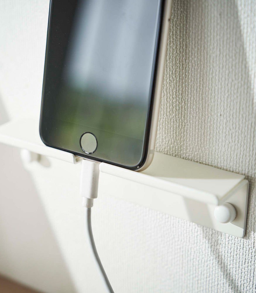 View 6 - An iPhone securely mounted on a wall, showcasing its screen while positioned vertically. The charger cord is neatly threaded through a designated opening in the mount, ensuring a streamlined appearance