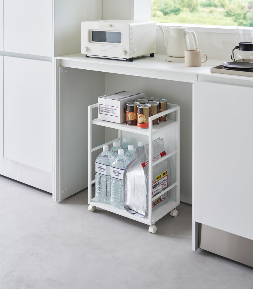 View 3 - Two-level white steel storage cart with casters is shown in an opening of a recessed cabinet. Cart houses a box and six cans of food on the upper shelf, the lower shelf holds a large bag of food with 6 liters of bottled water. One side of the cart has a wall to hide storage.