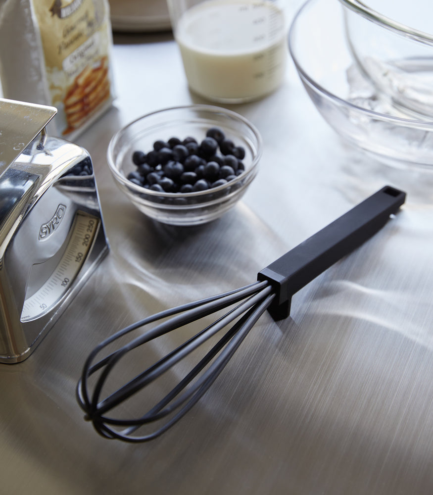 View 9 - A black Yamazaki Floating whisk resting on a table next to blueberries.