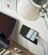 An iPhone with a charger cord plugged in is mounted on a wall above a desk setup. The screen is facing outward and the phone is held up on its side.