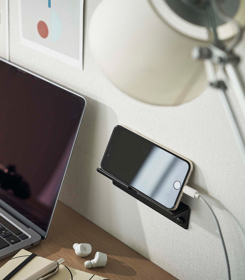 An iPhone with a charger cord plugged in is mounted on a wall above a desk setup. The screen is facing outward and the phone is held up on its side.