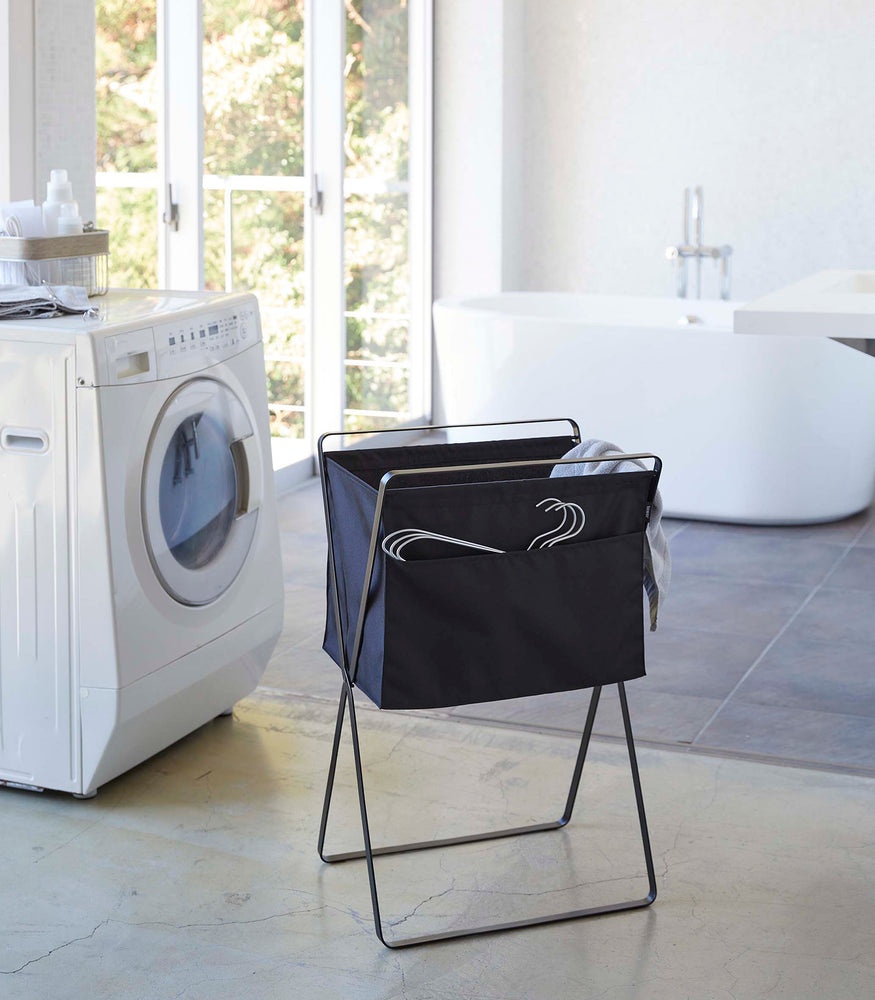 View 22 - A black laundry hamper with black metal legs is angled in front of a washing machine. Wired hangers are poking out of a pocket in the front of the hamper and a towel is seen inside. A washing machine and bathtub are visible in the background.