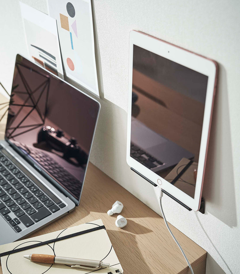A tablet is securely mounted on a wall, showcasing its screen while positioned vertically. The charger cord is neatly threaded through a designated opening in the mount, ensuring a streamlined appearance.